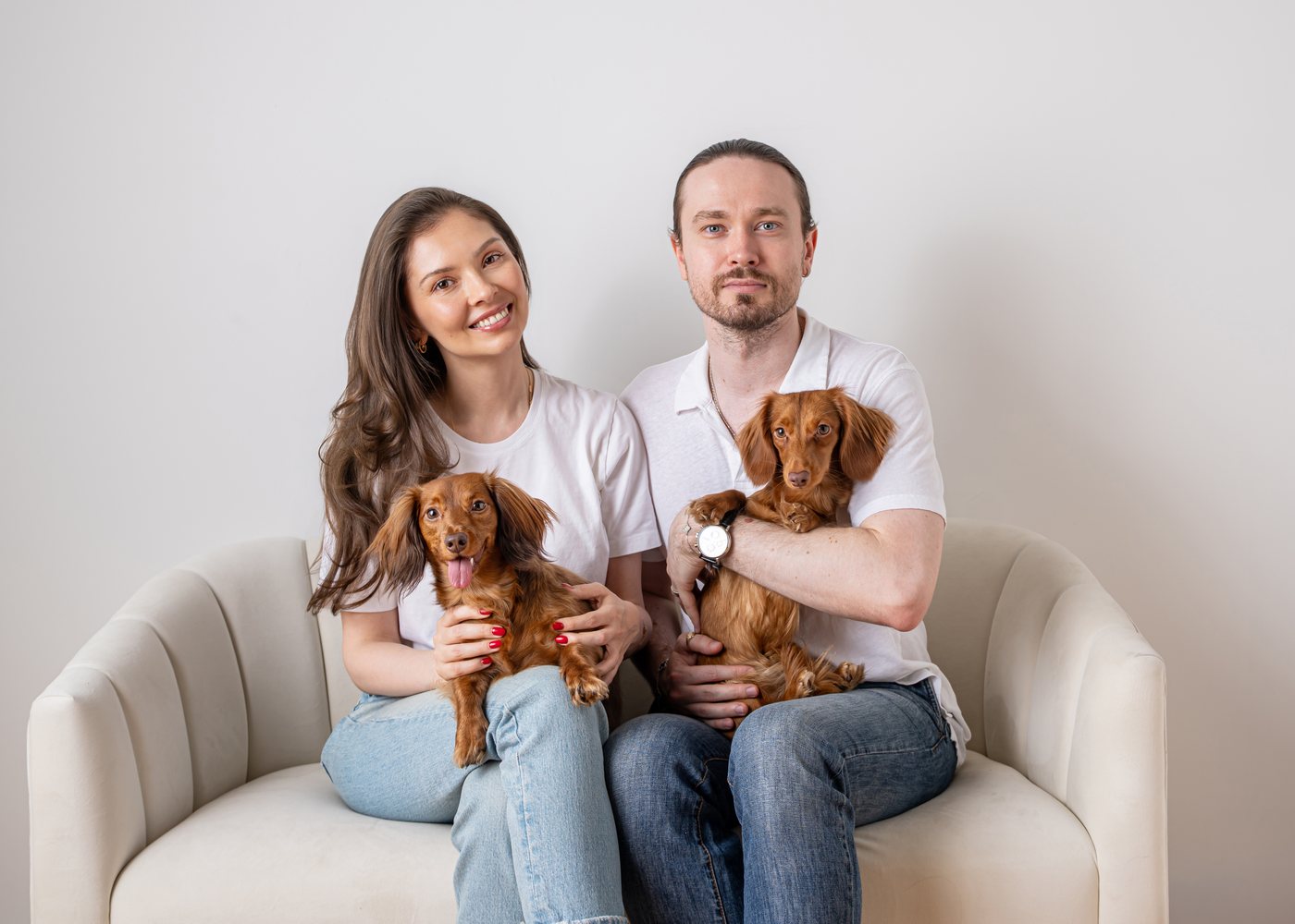 Iana and Alex, the founders of Nicole & Nora, sitting on a cream sofa holding their two long-haired mini dachshunds — Nicole on the left and Nora on the right